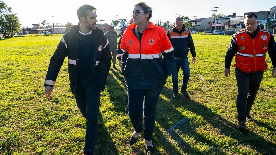 Foto mostra grupo de pessoas caminhando por um campo gramado, com Leite e Juliana à frente. Todos usam jaquetas com logotipos e emblemas de serviços de emergência, indicando que fazem parte de uma equipe de resgate ou atendimento. Ao fundo, há prédios, veículos e um helicóptero.