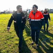 Foto mostra grupo de pessoas caminhando por um campo gramado, com Leite e Juliana à frente. Todos usam jaquetas com logotipos e emblemas de serviços de emergência, indicando que fazem parte de uma equipe de resgate ou atendimento. Ao fundo, há prédios, veículos e um helicóptero.