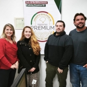 A foto mostra um grupo de cinco pessoas, três mulheres e dois homens, posando em pé na frente de um painel promocional do Programa Produtos Premium. O painel branco tem um logotipo no topo que diz "Selo de Reconhecimento Produtos Premium RS", com um ícone de "ORIGEM E QUALIDADE RS" e a palavra "PREMIUM" em destaque. À esquerda, as duas mulheres sorriem. A mulher do centro tem cabelos ruivos e usa uma jaqueta preta. À direita, os dois homens sorriem. O ambiente parece ser um corredor ou recepção, com paredes claras e portas no fundo. A cena sugere a concessão de um selo de qualidade para produtos gaúchos.