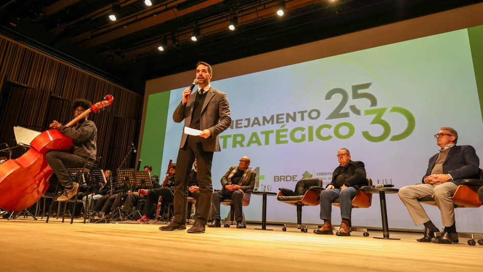 A foto mostra o governador Eduardo Leite de pé no palco, centralizado, discursando. Ele é um homem branco, vestindo um terno escuro, camisa branca e gravata preta. Leite segura um microfone com a mão esquerda e um papel com a direita, olhando para a plateia (não visível). Atrás dele, um grande telão branco exibe a frase "PLANEJAMENTO ESTRATÉGICO" e os números "25 30" em destaque. À esquerda do palco, um músico de cabelos crespos toca um violoncelo vermelho, com outros músicos parcialmente visíveis ao fundo, sugerindo uma orquestra. À direita de Leite, três homens adultos estão sentados em poltronas escuras. O palco é de madeira clara e o fundo apresenta cortinas escuras.