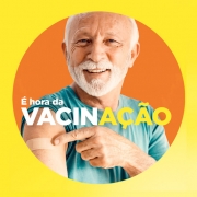 Card em fundo amarelo com um círculo em cor laranja ao centro. No círculo, há uma pessoa idosa de cabelo e barba branca, com camiseta azul. Ela aponta para um curativo no braço. A imagem traz o texto: “É hora da VACINAÇÃO”.