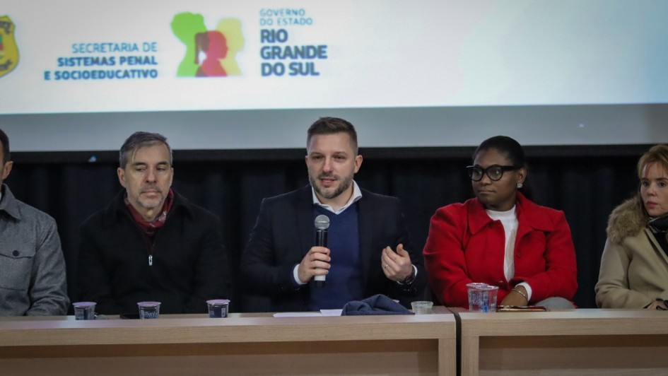 A foto mostra o secretário-adjunto da SSPS, Cesar Kurtz, um homem branco de barba e cabelo curto, sentado ao centro de uma mesa em um palco, falando ao microfone. Ele veste um blazer escuro sobre uma camisa azul e gesticula com a mão esquerda enquanto fala. Ao seu lado esquerdo, um homem de cabelo grisalho e casaco preto também está sentado. À direita de Cesar Kurtz, uma mulher negra de óculos e blazer vermelho está sentada, com as mãos sobre a mesa. Mais à direita, outra mulher loira de casaco claro e gola de pele está sentada. Ao fundo, um banner branco exibe o logotipo da "SECRETARIA DE SISTEMAS PENAL E SOCIOEDUCATIVO" e o brasão do "GOVERNO DO ESTADO RIO GRANDE DO SUL", com um ícone colorido de figuras humanas. A cena é de um seminário ou evento público.