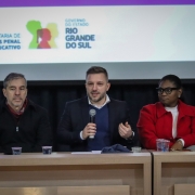 A foto mostra o secretário-adjunto da SSPS, Cesar Kurtz, um homem branco de barba e cabelo curto, sentado ao centro de uma mesa em um palco, falando ao microfone. Ele veste um blazer escuro sobre uma camisa azul e gesticula com a mão esquerda enquanto fala. Ao seu lado esquerdo, um homem de cabelo grisalho e casaco preto também está sentado. À direita de Cesar Kurtz, uma mulher negra de óculos e blazer vermelho está sentada, com as mãos sobre a mesa. Mais à direita, outra mulher loira de casaco claro e gola de pele está sentada. Ao fundo, um banner branco exibe o logotipo da "SECRETARIA DE SISTEMAS PENAL E SOCIOEDUCATIVO" e o brasão do "GOVERNO DO ESTADO RIO GRANDE DO SUL", com um ícone colorido de figuras humanas. A cena é de um seminário ou evento público.