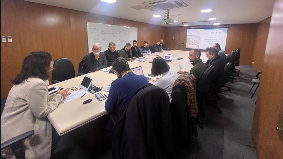 Na foto, há um grupo de pessoas sentadas ao redor de uma grande mesa de conferência em uma sala de reuniões. As paredes são de madeira, há um quadro branco com anotações e uma tela de projetor exibindo uma apresentação. Sobre a mesa, estão espalhados notebooks, cadernos e outros materiais de trabalho.