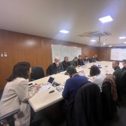 Na foto, há um grupo de pessoas sentadas ao redor de uma grande mesa de conferência em uma sala de reuniões. As paredes são de madeira, há um quadro branco com anotações e uma tela de projetor exibindo uma apresentação. Sobre a mesa, estão espalhados notebooks, cadernos e outros materiais de trabalho.