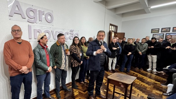 A foto mostra um grupo de pessoas em um ambiente interno, que parece ser um salão ou auditório, com paredes brancas e um piso de madeira com padrão em espinha de peixe. À esquerda, em destaque na parede, há letras grandes em relevo que formam "Agro Inovação". No centro da imagem, está o secretário da Agricultura, Edivilson Brum, um homem de colete azul marinho, camisa azul clara e calça jeans escura, está de pé segurando um microfone e gesticulando com a mão direita, olhando para a plateia (parcialmente visível à direita). Várias pessoas, em sua maioria homens, estão de pé e sentadas ao redor, observando o orador. A cena é um evento ou reunião relacionada à agricultura.