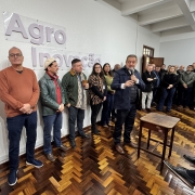 A foto mostra um grupo de pessoas em um ambiente interno, que parece ser um salão ou auditório, com paredes brancas e um piso de madeira com padrão em espinha de peixe. À esquerda, em destaque na parede, há letras grandes em relevo que formam "Agro Inovação". No centro da imagem, está o secretário da Agricultura, Edivilson Brum, um homem de colete azul marinho, camisa azul clara e calça jeans escura, está de pé segurando um microfone e gesticulando com a mão direita, olhando para a plateia (parcialmente visível à direita). Várias pessoas, em sua maioria homens, estão de pé e sentadas ao redor, observando o orador. A cena é um evento ou reunião relacionada à agricultura.