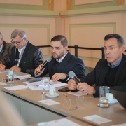 Foto de cinco homens sentados lado a lado em uma mesa de reunião. Todos usam trajes formais. Gabriel, ao centro, segura um microfone e fala; à sua frente há papéis, um copo com água e uma xícara.