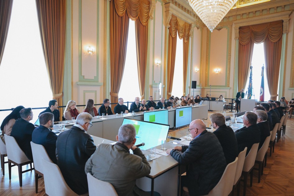 Foto da reunião em uma sala de estilo clássico, com lustre de cristal, cortinas douradas e paredes decoradas. Mais de 30 pessoas estão sentadas ao redor de uma mesa retangular, participando da discussão. Três grandes telas mostram uma apresentação digital. Ao fundo, há bandeiras oficiais e uma pessoa em pé ao lado de um púlpito.