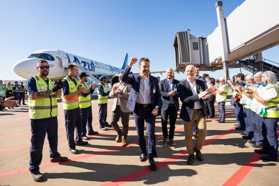 A imagem mostra o governador do Rio Grande do Sul, Eduardo Leite, e o CEO da Fraport, Stefan Schulte, caminhando em uma pista de aeroporto, sendo aplaudidos por funcionários em ambos os lados. Ao fundo, à esquerda, há um avião da Azul Linhas Aéreas. O céu está claro e azul. A cena parece ser a reabertura ou um evento importante no Aeroporto Salgado Filho, em Porto Alegre, dado o contexto da imagem.