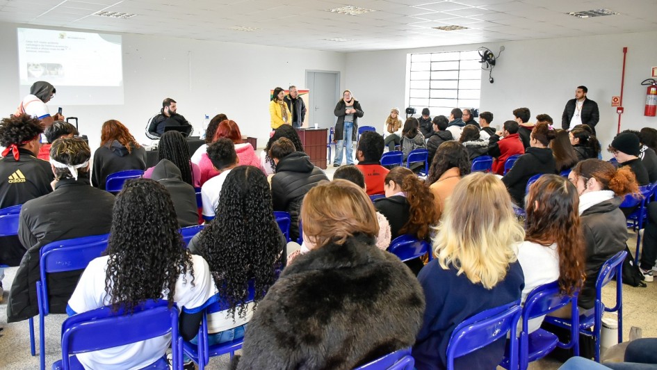 Uma sala de paredes brancas com fileiras de cadeiras azuis. Pessoas estão sentadas, voltadas para a frente, onde cinco apresentadores estão em pé diante de uma tela de projeção exibindo slides. A plateia acompanha atentamente a apresentação em um ambiente iluminado e formal.