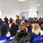 Uma sala de paredes brancas com fileiras de cadeiras azuis. Pessoas estão sentadas, voltadas para a frente, onde cinco apresentadores estão em pé diante de uma tela de projeção exibindo slides. A plateia acompanha atentamente a apresentação em um ambiente iluminado e formal.