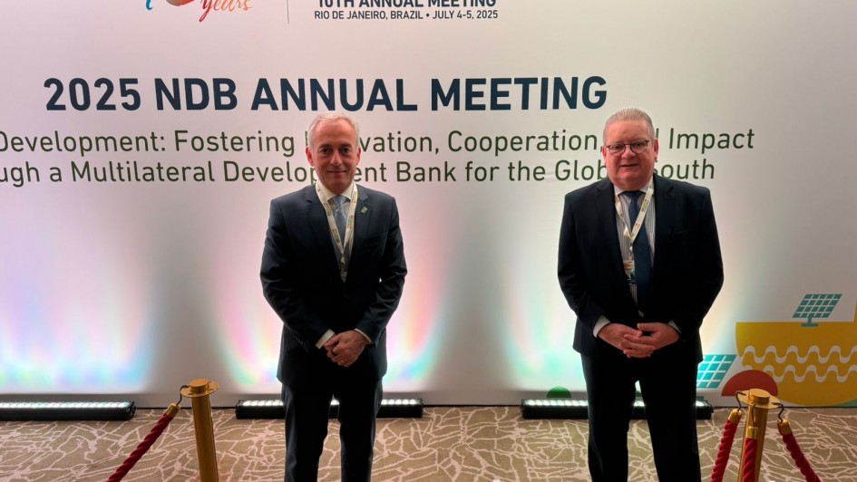 A imagem mostra duas pessoas, aparentemente homens, de pé e sorrindo para a câmera em frente a um grande banner. Ambos estão usando ternos escuros. O banner, ao fundo, é predominantemente branco e exibe o logotipo do "New Development Bank" com a inscrição "10 years" (10 anos), e um cabeçalho vermelho com "NDB2025 - 10TH ANNUAL MEETING - RIO DE JANEIRO, BRAZIL - JULY 4-5, 2025". Abaixo, em letras grandes, está escrito "2025 NDB ANNUAL MEETING", seguido de um texto menor que parece ser o tema do evento: "Development: Fostering Innovation, Cooperation, and Impact through a Multilateral Development Bank for the Global South". Há um pequeno ícone de painéis solares no canto inferior direito do banner. Duas cordas de isolamento douradas com suportes escuros marcam o espaço à frente das pessoas.
