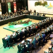 Foto mostra uma grande reunião em formato de mesa em U, com dezenas de pessoas sentadas ao redor, todas usando notebooks e microfones. No centro do salão, há uma decoração com elementos da floresta e frutas típicas da Amazônia, iluminados com luz verde. Ao fundo, vê-se um grande painel escrito “COMSEFAZ – CONFAZ”.