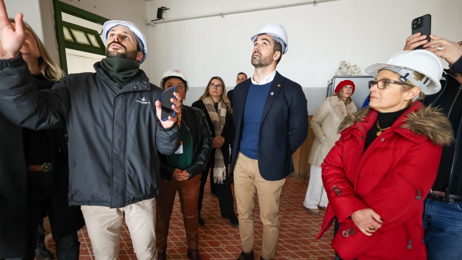 Foto mostra várias pessoas numa sala fechada, todas de capacete de proteção. À esquerda, um homem aponta algo, para onde as pessoas olham. Leite está ao centro, em pé, olhando ligeiramente para o alto.
