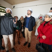 Foto mostra várias pessoas numa sala fechada, todas de capacete de proteção. À esquerda, um homem aponta algo, para onde as pessoas olham. Leite está ao centro, em pé, olhando ligeiramente para o alto.