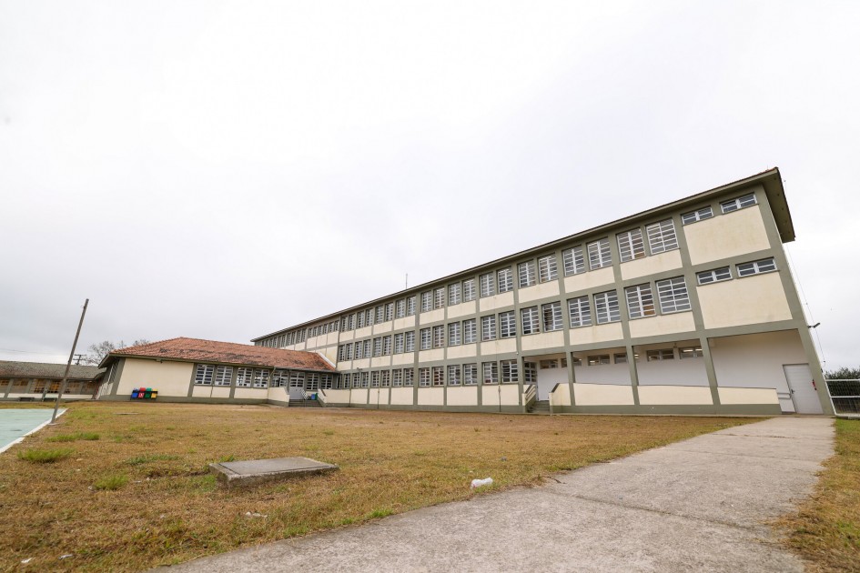 Foto de prédio escolar grande, de três andares, com muitas janelas enfileiradas. O dia está nublado. À frente, um pátio gramado com uma calçada de concreto leva até o prédio.