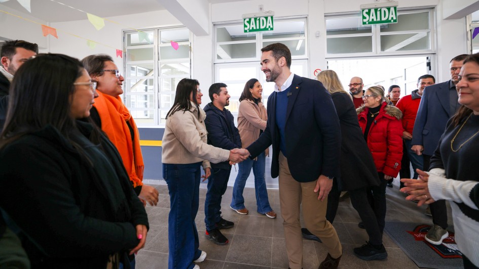 Foto de Leite usando blazer escuro e calça bege cumprimentando pessoas na entrada da escola. Há um grupo de pessoas sorrindo, e bandeirinhas de festa junina decoram o espaço.