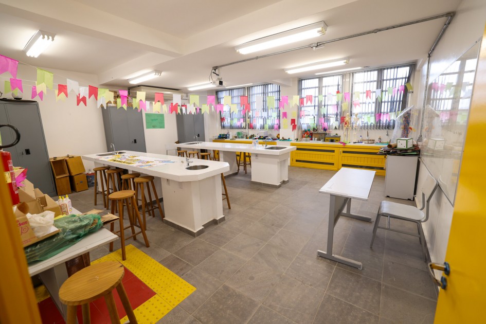 Foto de sala ampla com bancadas brancas, banquetas de madeira e materiais didáticos ao fundo. Bandeirinhas coloridas decoram o teto. A sala está organizada e iluminada, com elementos de acessibilidade no chão.