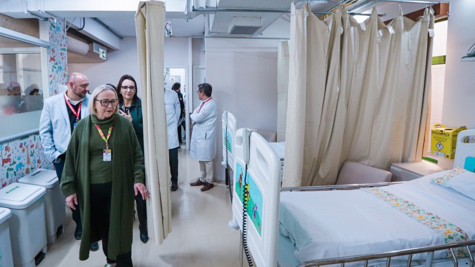 A imagem mostra o interior de um quarto de hospital espaçoso, com paredes brancas e claras. À direita, há duas camas de hospital vazias, arrumadas com lençóis brancos e travesseiros, separadas por cortinas bege. A cama mais próxima da câmera tem um lençol com desenhos coloridos na parte de baixo.
No centro da imagem, uma mulher loira de meia-idade, usando um casaco verde escuro comprido e calças pretas, caminha em direção ao observador. Ao fundo, outras pessoas, incluindo profissionais de saúde em jalecos brancos, estão de pé e conversando. No lado esquerdo, é possível ver um cesto de lixo branco e uma parede decorada com adesivos coloridos. A iluminação é clara e uniforme, sugerindo um ambiente bem cuidado e limpo.