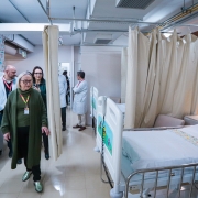 A imagem mostra o interior de um quarto de hospital espaçoso, com paredes brancas e claras. À direita, há duas camas de hospital vazias, arrumadas com lençóis brancos e travesseiros, separadas por cortinas bege. A cama mais próxima da câmera tem um lençol com desenhos coloridos na parte de baixo.

No centro da imagem, uma mulher loira de meia-idade, usando um casaco verde escuro comprido e calças pretas, caminha em direção ao observador. Ao fundo, outras pessoas, incluindo profissionais de saúde em jalecos brancos, estão de pé e conversando. No lado esquerdo, é possível ver um cesto de lixo branco e uma parede decorada com adesivos coloridos. A iluminação é clara e uniforme, sugerindo um ambiente bem cuidado e limpo.