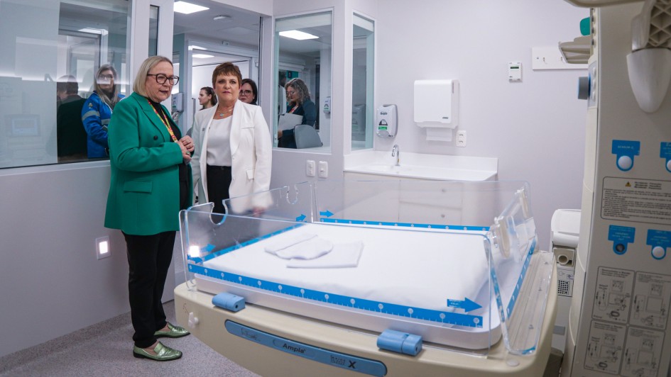 A imagem mostra uma sala de hospital, possivelmente uma unidade neonatal, com um berço aquecido em primeiro plano. Duas pessoas estão conversando ao lado do berço. Arita Bergmann está vestida com um blazer verde e a outra com um blazer branco. Ao fundo, há mais pessoas observando através de uma janela de vidro. A sala é bem iluminada e possui equipamentos médicos nas paredes e no teto.