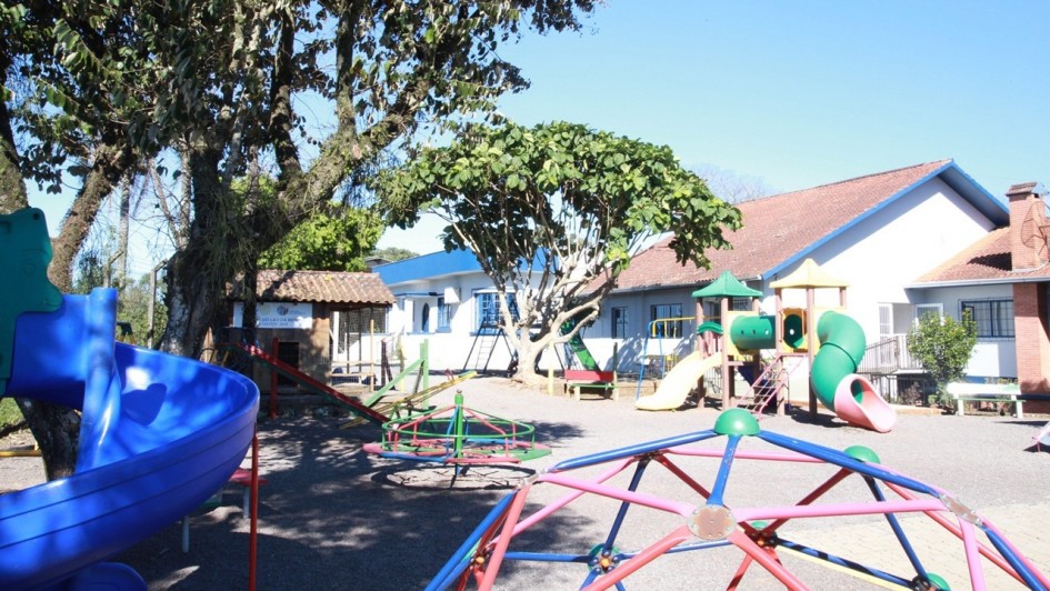 A foto mostra um playground externo em um dia ensolarado. No primeiro plano, à esquerda, uma estrutura de escorregador azul brilhante com detalhes vermelhos domina o lado esquerdo da imagem. No centro e à direita, uma série de brinquedos de playground coloridos, incluindo escorregadores amarelos, verdes e cor-de-rosa, um carrossel vermelho e verde, e uma estrutura de escalada geométrica azul e vermelha, estão dispostos sobre um piso de cascalho cinza. Atrás do playground, há um edifício branco de um andar com telhados de telha marrom-avermelhada e detalhes em azul, sugerindo uma escola ou creche. Duas árvores grandes e frondosas, com folhagem verde escura, fornecem sombra à esquerda e no centro da imagem. O céu é azul claro, sem nuvens.