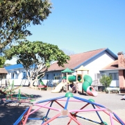 A foto mostra um playground externo em um dia ensolarado. No primeiro plano, à esquerda, uma estrutura de escorregador azul brilhante com detalhes vermelhos domina o lado esquerdo da imagem. No centro e à direita, uma série de brinquedos de playground coloridos, incluindo escorregadores amarelos, verdes e cor-de-rosa, um carrossel vermelho e verde, e uma estrutura de escalada geométrica azul e vermelha, estão dispostos sobre um piso de cascalho cinza. Atrás do playground, há um edifício branco de um andar com telhados de telha marrom-avermelhada e detalhes em azul, sugerindo uma escola ou creche. Duas árvores grandes e frondosas, com folhagem verde escura, fornecem sombra à esquerda e no centro da imagem. O céu é azul claro, sem nuvens.