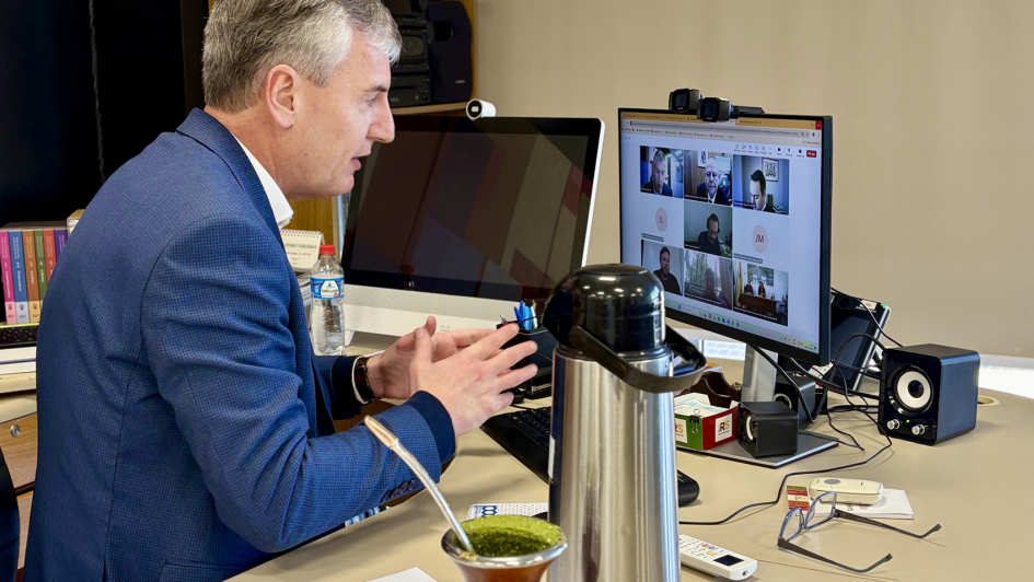 O Secretário da Cultura do RS, Eduardo Loureiro, aparece na foto. Ele é um homem de meia-idade, com cabelos curtos e grisalhos, está sentado em uma mesa de escritório, voltado para a esquerda e olhando para um monitor de computador. Ele veste um terno azul-marinho sobre uma camisa branca e gesticula com as mãos abertas enquanto fala. À sua frente, sobre a mesa bege, há um monitor de computador à esquerda exibindo uma imagem escura, e um monitor maior à direita, mostrando uma videochamada com quatro participantes em pequenas janelas.
Entre os monitores e o homem, repousam uma cuia de chimarrão com a bomba e um recipiente térmico prateado. Há também um mouse, óculos, um controle remoto, caixas de som e papéis espalhados pela mesa. Ao fundo, uma estante de madeira com pastas e objetos diversos pode ser vista. A iluminação é suave, vinda de cima e da frente.