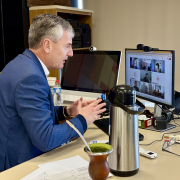O Secretário da Cultura do RS, Eduardo Loureiro, aparece na foto. Ele é um homem de meia-idade, com cabelos curtos e grisalhos, está sentado em uma mesa de escritório, voltado para a esquerda e olhando para um monitor de computador. Ele veste um terno azul-marinho sobre uma camisa branca e gesticula com as mãos abertas enquanto fala. À sua frente, sobre a mesa bege, há um monitor de computador à esquerda exibindo uma imagem escura, e um monitor maior à direita, mostrando uma videochamada com quatro participantes em pequenas janelas.
Entre os monitores e o homem, repousam uma cuia de chimarrão com a bomba e um recipiente térmico prateado. Há também um mouse, óculos, um controle remoto, caixas de som e papéis espalhados pela mesa. Ao fundo, uma estante de madeira com pastas e objetos diversos pode ser vista. A iluminação é suave, vinda de cima e da frente.
