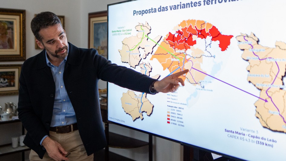 A imagem mostra um homem, identificado como o governador Eduardo Leite, de pé, apontando para uma grande tela de apresentação. Ele veste uma camisa azul clara, um blazer escuro e calças cáqui. A tela exibe um mapa com "Proposta das variantes ferroviárias" no topo. O mapa é estilizado com diferentes tons de marrom, laranja e vermelho, indicando áreas ou rotas, e algumas linhas verdes e roxas representando as variantes ferroviárias. Há texto e legendas na tela, incluindo detalhes sobre as variantes e "CAPEX". Ao fundo, à esquerda, há um ambiente interno com quadros emoldurados na parede e um bule de chá sobre um móvel.