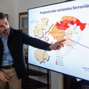 A imagem mostra um homem, identificado como o governador Eduardo Leite, de pé, apontando para uma grande tela de apresentação. Ele veste uma camisa azul clara, um blazer escuro e calças cáqui. A tela exibe um mapa com "Proposta das variantes ferroviárias" no topo. O mapa é estilizado com diferentes tons de marrom, laranja e vermelho, indicando áreas ou rotas, e algumas linhas verdes e roxas representando as variantes ferroviárias. Há texto e legendas na tela, incluindo detalhes sobre as variantes e "CAPEX". Ao fundo, à esquerda, há um ambiente interno com quadros emoldurados na parede e um bule de chá sobre um móvel.