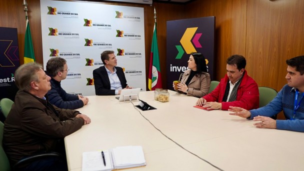 Uma fotografia captura uma reunião em uma sala de conferências, com seis pessoas sentadas ao redor de uma mesa retangular clara. No centro da imagem, em frente a um painel branco com múltiplos logotipos "Rio Grande do Sul" e folhas estilizadas em tons de verde e marrom, está o Secretário de Desenvolvimento Econômico, Ernani Polo. Ele veste um terno escuro e uma camisa clara, e está olhando para a direita, engajado na conversa.
À esquerda de Polo, dois homens estão sentados, um com um casaco azul-marinho e outro com um casaco marrom, ambos olhando na direção do secretário.
À direita de Polo, uma mulher de cabelos escuros e casaco bege segura uma xícara, olhando para os participantes à sua direita. Ao lado dela, um homem com um casaco vermelho e camisa branca gesticula com a mão esquerda, enquanto o último homem à direita, com uma camisa azul, também gesticula enquanto olha para a frente.
. No canto superior direito, um painel escuro com um logotipo vibrante "Invest" em tons de roxo, amarelo e verde está visível, e duas bandeiras, uma do Brasil e outra do Rio Grande do Sul, ladeiam o painel principal na parede. A sala tem paredes de madeira e um teto com iluminação embutida.