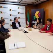 Uma fotografia captura uma reunião em uma sala de conferências, com seis pessoas sentadas ao redor de uma mesa retangular clara. No centro da imagem, em frente a um painel branco com múltiplos logotipos "Rio Grande do Sul" e folhas estilizadas em tons de verde e marrom, está o Secretário de Desenvolvimento Econômico, Ernani Polo. Ele veste um terno escuro e uma camisa clara, e está olhando para a direita, engajado na conversa.
À esquerda de Polo, dois homens estão sentados, um com um casaco azul-marinho e outro com um casaco marrom, ambos olhando na direção do secretário.
À direita de Polo, uma mulher de cabelos escuros e casaco bege segura uma xícara, olhando para os participantes à sua direita. Ao lado dela, um homem com um casaco vermelho e camisa branca gesticula com a mão esquerda, enquanto o último homem à direita, com uma camisa azul, também gesticula enquanto olha para a frente.
. No canto superior direito, um painel escuro com um logotipo vibrante "Invest" em tons de roxo, amarelo e verde está visível, e duas bandeiras, uma do Brasil e outra do Rio Grande do Sul, ladeiam o painel principal na parede. A sala tem paredes de madeira e um teto com iluminação embutida.