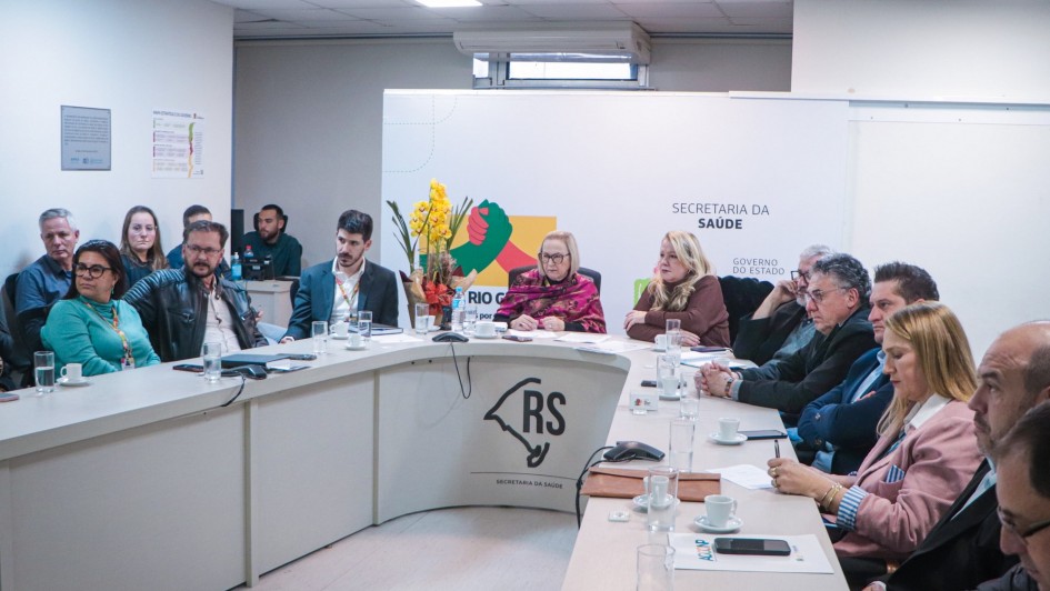 A imagem mostra uma reunião em uma sala moderna, com um grupo de pessoas sentadas ao redor de uma mesa em formato de "U". A maioria dos participantes é adulta, e estão vestidos de forma formal ou semi-formal. No fundo da sala, há uma parede branca com a logomarca da Secretaria da Saúde do Governo do Estado do Rio Grande do Sul e a inscrição "SECRETARIA DA SAÚDE" em destaque. À frente da mesa, há uma bandeira estilizada do Rio Grande do Sul com a sigla "RS". Há xícaras de café e outros objetos sobre a mesa.
