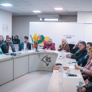 A imagem mostra uma reunião em uma sala moderna, com um grupo de pessoas sentadas ao redor de uma mesa em formato de "U". A maioria dos participantes é adulta, e estão vestidos de forma formal ou semi-formal. No fundo da sala, há uma parede branca com a logomarca da Secretaria da Saúde do Governo do Estado do Rio Grande do Sul e a inscrição "SECRETARIA DA SAÚDE" em destaque. À frente da mesa, há uma bandeira estilizada do Rio Grande do Sul com a sigla "RS". Há xícaras de café e outros objetos sobre a mesa.