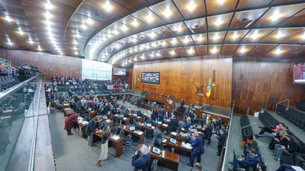 A imagem é uma vista panorâmica de um plenário de uma Assembleia Legislativa, provavelmente a do Rio Grande do Sul, visto de um ângulo elevado. O teto é de madeira em tom escuro, com iluminação embutida. No centro, há uma grande área com mesas e cadeiras dispostas em semicírculo, onde deputados e funcionários estão sentados, alguns usando laptops. Ao fundo, no palco principal, há uma bancada com microfones e bandeiras (provavelmente do Brasil e do Rio Grande do Sul), além de um painel eletrônico e revestimento de madeira. Nas laterais e na parte superior, há galerias com assentos para o público, onde algumas pessoas estão sentadas. A sala está bem iluminada.
