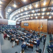 A imagem é uma vista panorâmica de um plenário de uma Assembleia Legislativa, provavelmente a do Rio Grande do Sul, visto de um ângulo elevado. O teto é de madeira em tom escuro, com iluminação embutida. No centro, há uma grande área com mesas e cadeiras dispostas em semicírculo, onde deputados e funcionários estão sentados, alguns usando laptops. Ao fundo, no palco principal, há uma bancada com microfones e bandeiras (provavelmente do Brasil e do Rio Grande do Sul), além de um painel eletrônico e revestimento de madeira. Nas laterais e na parte superior, há galerias com assentos para o público, onde algumas pessoas estão sentadas. A sala está bem iluminada.