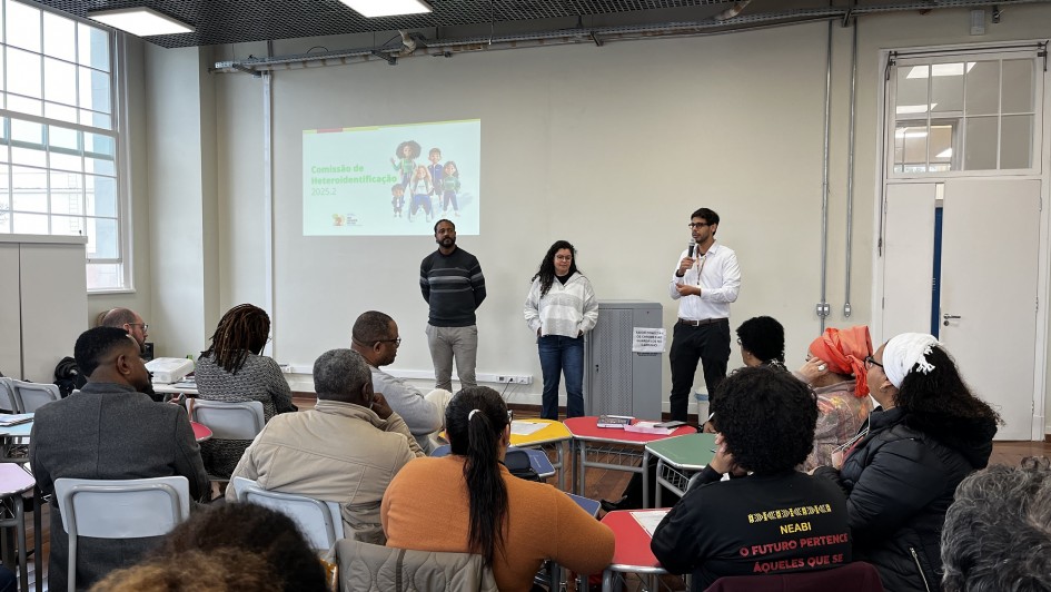 A imagem mostra uma sala de aula ou auditório onde um grupo de pessoas está assistindo a uma apresentação. No palco ou à frente da sala, três pessoas (dois homens e uma mulher) estão de pé. Um dos homens, à direita, está falando em um microfone. Atrás deles, há uma tela de projeção com uma apresentação visual, que inclui a frase "Comissão de Heteroidentificação". A plateia é composta por pessoas sentadas em carteiras ou cadeiras, de costas ou de perfil, observando os apresentadores. A sala tem grandes janelas e iluminação no teto.
