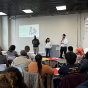 A imagem mostra uma sala de aula ou auditório onde um grupo de pessoas está assistindo a uma apresentação. No palco ou à frente da sala, três pessoas (dois homens e uma mulher) estão de pé. Um dos homens, à direita, está falando em um microfone. Atrás deles, há uma tela de projeção com uma apresentação visual, que inclui a frase "Comissão de Heteroidentificação". A plateia é composta por pessoas sentadas em carteiras ou cadeiras, de costas ou de perfil, observando os apresentadores. A sala tem grandes janelas e iluminação no teto.