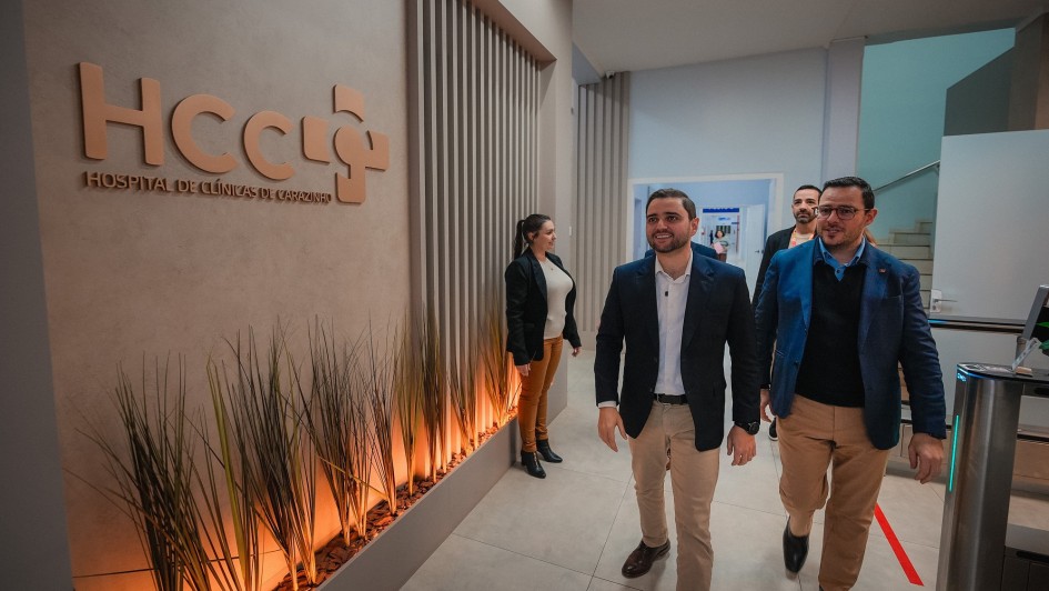 A imagem mostra um grupo de pessoas caminhando por um corredor moderno dentro de um hospital. Em primeiro plano, dois homens estão andando lado a lado, sorrindo e olhando para a direita. O homem à frente veste um blazer escuro, camisa branca e calças cáqui. O outro homem, ao lado dele, veste um blazer azul, camisa azul e calças cáqui. À esquerda, em destaque na parede, há uma placa com o logo "HCCI" em letras grandes e, abaixo, "HOSPITAL DE CLÍNICAS DE CARAZINHO". A parede tem detalhes verticais amadeirados e, na parte inferior, há uma jardineira com plantas. Mais ao fundo, outras pessoas são visíveis no corredor.
