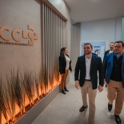 A imagem mostra um grupo de pessoas caminhando por um corredor moderno dentro de um hospital. Em primeiro plano, dois homens estão andando lado a lado, sorrindo e olhando para a direita. O homem à frente veste um blazer escuro, camisa branca e calças cáqui. O outro homem, ao lado dele, veste um blazer azul, camisa azul e calças cáqui. À esquerda, em destaque na parede, há uma placa com o logo "HCCI" em letras grandes e, abaixo, "HOSPITAL DE CLÍNICAS DE CARAZINHO". A parede tem detalhes verticais amadeirados e, na parte inferior, há uma jardineira com plantas. Mais ao fundo, outras pessoas são visíveis no corredor.