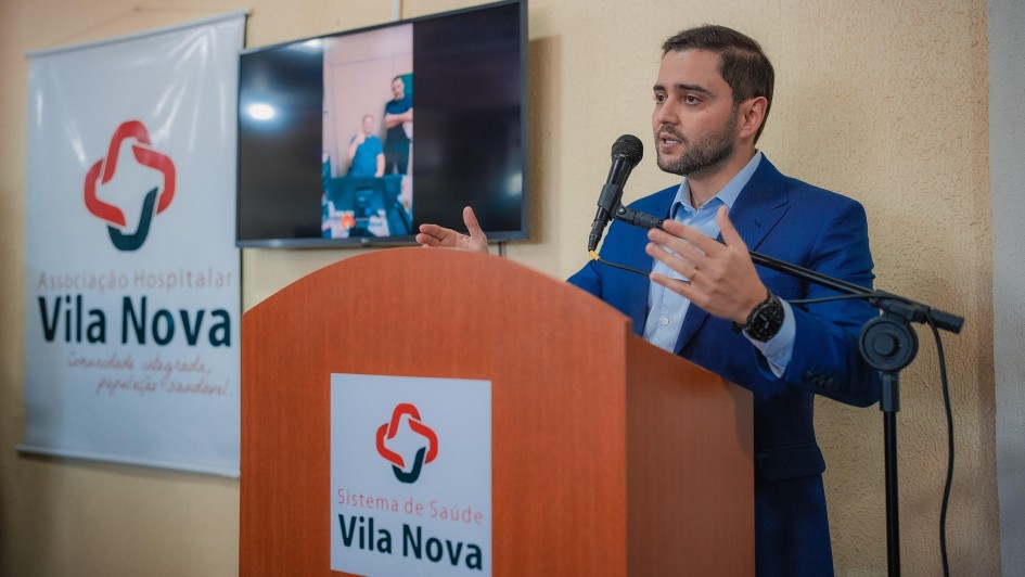 A foto, em ambiente interno, mostra o vice-governador Gabriel Souza, um homem de pele clara e barba, falando em um púlpito de madeira. Ele veste um terno azul xadrez e uma camisa azul clara, e gesticula com as mãos enquanto fala ao microfone. No púlpito, há o logotipo vermelho e branco de um coração e uma cruz, e as palavras "Sistema de Saúde Vila Nova" em preto. À esquerda do púlpito, um banner branco exibe o mesmo logotipo e as palavras "Associação Hospitalar Vila Nova" em vermelho e preto. Atrás do vice-governador, uma televisão de tela plana exibe uma imagem de duas pessoas em desfoque. O fundo é uma parede bege simples. A iluminação geral é clara.