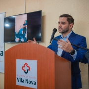 A foto, em ambiente interno, mostra o vice-governador Gabriel Souza, um homem de pele clara e barba, falando em um púlpito de madeira. Ele veste um terno azul xadrez e uma camisa azul clara, e gesticula com as mãos enquanto fala ao microfone. No púlpito, há o logotipo vermelho e branco de um coração e uma cruz, e as palavras "Sistema de Saúde Vila Nova" em preto. À esquerda do púlpito, um banner branco exibe o mesmo logotipo e as palavras "Associação Hospitalar Vila Nova" em vermelho e preto. Atrás do vice-governador, uma televisão de tela plana exibe uma imagem de duas pessoas em desfoque. O fundo é uma parede bege simples. A iluminação geral é clara.