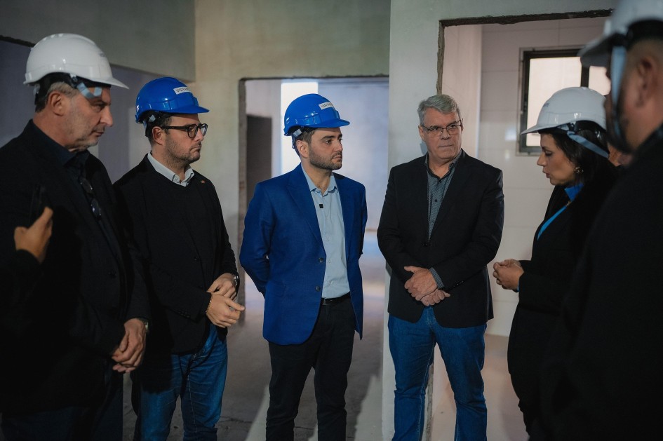 A fotografia mostra um grupo de seis pessoas em um ambiente interno que parece ser uma construção ou reforma. Todos usam capacetes de segurança (brancos ou azuis). No centro da imagem, o vice-governador Gabriel Souza, de capacete azul e terno azul, olha para a direita. Ao seu lado direito, um homem de óculos e capacete azul e outro de capacete branco. A mulher do grupo está à direita, de cabelos escuros e capacete branco, conversando com o homem ao seu lado. O ambiente é de concreto aparente e paredes em construção ou reforma, com uma janela visível ao fundo. A iluminação é difusa.