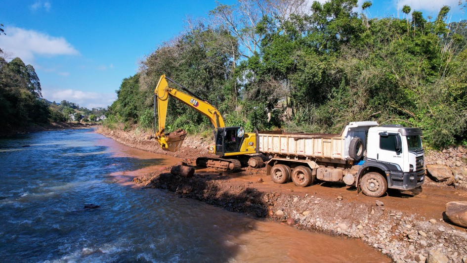 A imagem mostra uma escavadeira em operação em um rio, transferindo terra para a carroceria de um caminhão branco. Ambos os veículos estão posicionados sobre uma superfície de terra ao lado do rio.