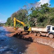A imagem mostra uma escavadeira em operação em um rio, transferindo terra para a carroceria de um caminhão branco. Ambos os veículos estão posicionados sobre uma superfície de terra ao lado do rio.