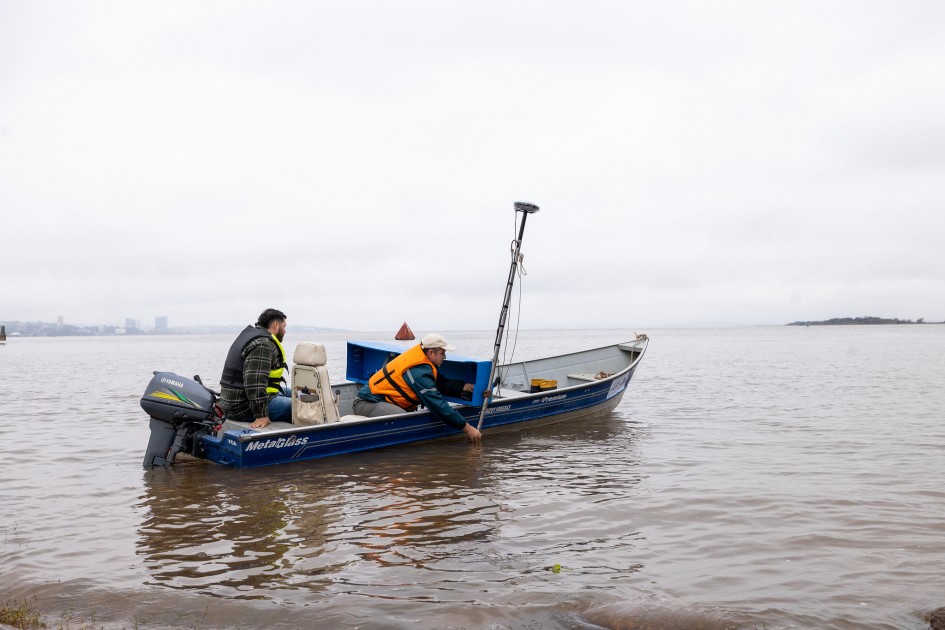 Dois homens estão em um pequeno barco no Guaíba, um veste colete salva-vidas laranja e está debruçado para fora do barco, manuseando equipamento com sonar e GPS. O outro está sentado, usando um colete preto e amarelo, observando a ação.