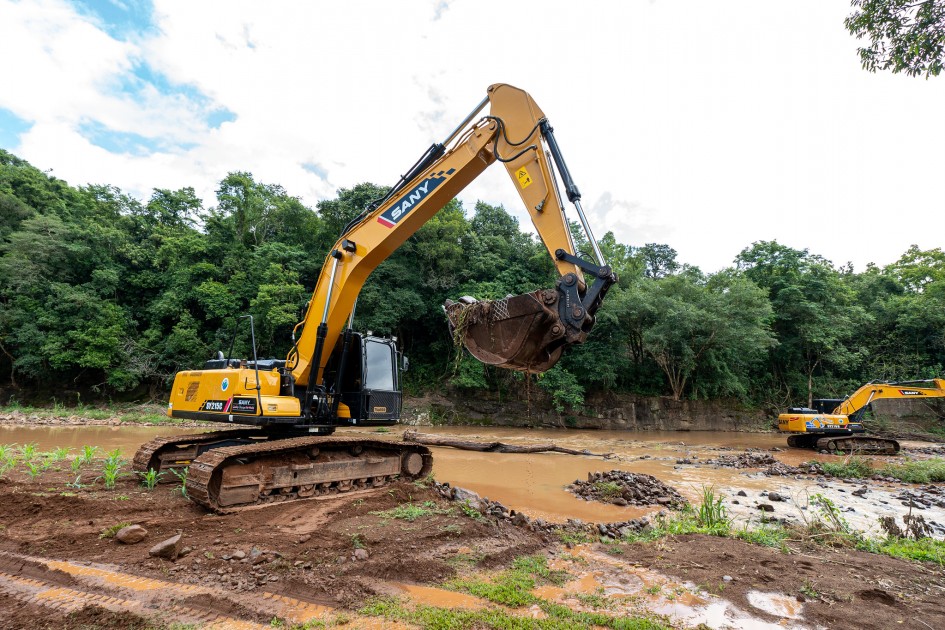 Duas escavadeiras amarelas estão em pleno funcionamento, realizando movimentação de terra para desassorear um leito de rio. A escavadeira em primeiro plano tem sua pá erguida, carregando uma mistura de terra e vegetação. Ao fundo, a segunda escavadeira está mais próxima de um riacho com água barrenta, cercado por pedras irregulares.