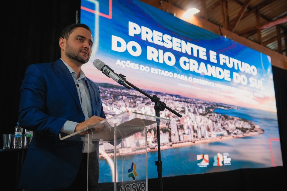 A foto mostra o vice-governador Gabriel Souza, de pele clara e barba, falando em um púlpito transparente com um microfone. Ele usa um terno azul-marinho e camisa clara, olhando para a esquerda da imagem. Atrás dele, um grande telão exibe a frase em letras maiúsculas "PRESENTE E FUTURO DO RIO GRANDE DO SUL" e abaixo "AÇÕES DO ESTADO PARA O DESENVOLVIMENTO...". A imagem de fundo no telão é uma vista aérea noturna de uma cidade litorânea com muitas luzes acesas, e um lago ou mar. O ambiente tem um teto de madeira e iluminação difusa.